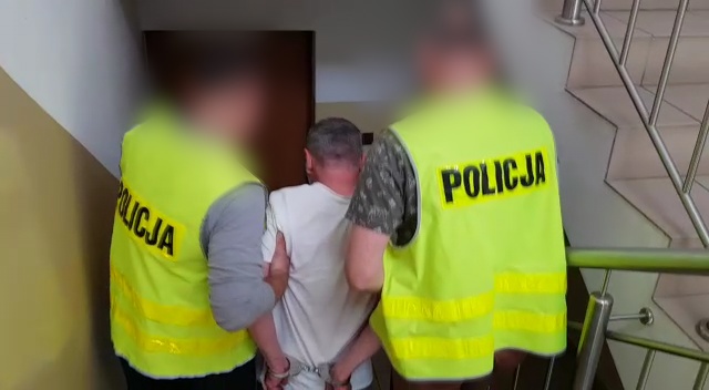 Policjanci ubrani w kamizelki z napisem policja prowadzą zatrzymanego po schodach w Komendzie Powiatowej Policji w Puławach.