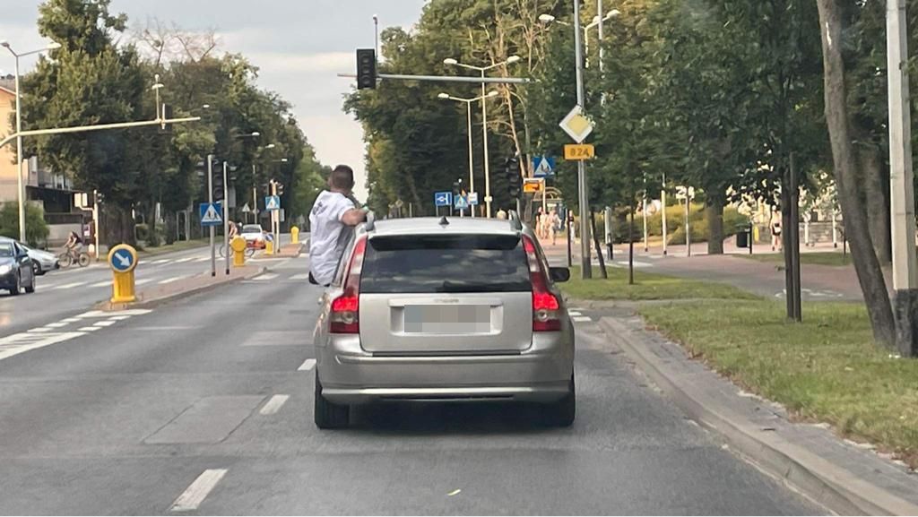 Na zdjęciu widzimy mężczyznę, który w czasie jazdy wysiadł zza kierownicy auta na zewnątrz pojazdu.