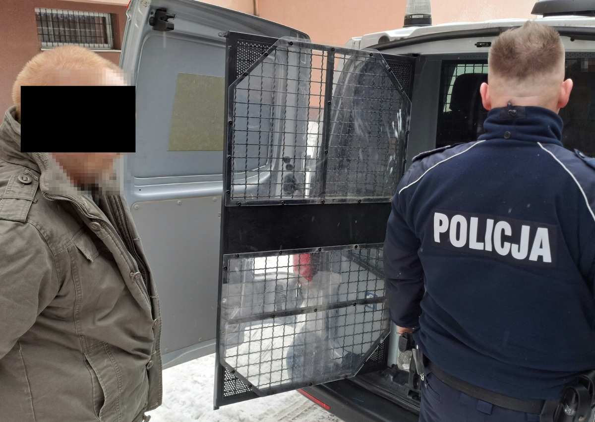 Zatrzymany mężczyzna wsiadający do okratowanego busa przy którym stoi policjant