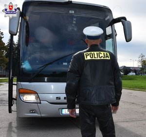 Policjant ruchu drogowego w białej czapce stoi przed szarym autokarem sprawdzając światła