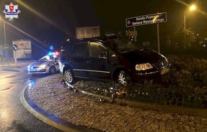 czarny volkswagen stojący na środku ronda wśród roślin, w tle radiowóz