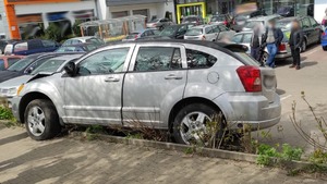 szary samochód stojący częściowo na chodniku a częściowo na skarpie między parkingiem a chodnikiem w tle inne samochody