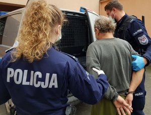 policjantka i policjant prowadzący zatrzymanego mężczyznę w kajdankach przed busem z kratą