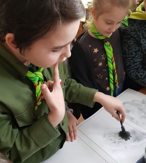 dzieci stojące przy biurku wpatrzone w kartkę na której chłopiec maluje pędzelkiem