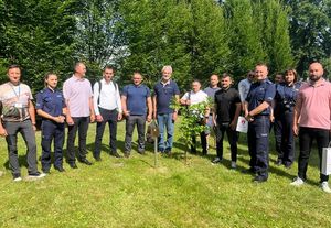 grupa ludzi oraz troje policjantów stojących na trawie przed nimi młody dąb