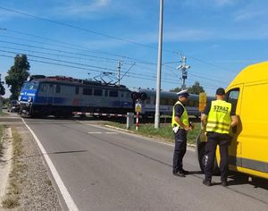 policjant i funkcjonariusz sok obok busa przed przejazdem, po torach jedzie pociąg