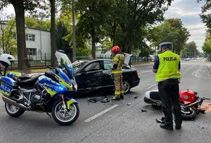 policjant i strażak na miejscu wypadku drogowego na ulicy leży motocykl stoi samochód mercedes oraz motocykl policyjny