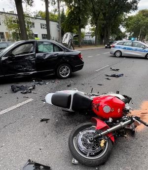 czerwono biały rozbity motocykl na jezdni w tle mercedes z rozbitym bokiem i radiowóz blokujący drogę