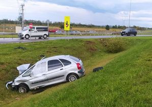 srebrny rozbity samochód osobowy w rowie, w tle dwa inne samochody jadące drogą