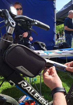 dłonie naklejające naklejkę na oznakowany rower w tle policjantka za stołem z ulotkami