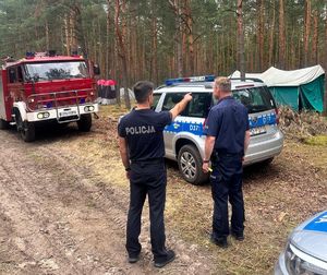 dwóch policjantów obok radiowozów w tle namioty harcerzy i wóz strażacki