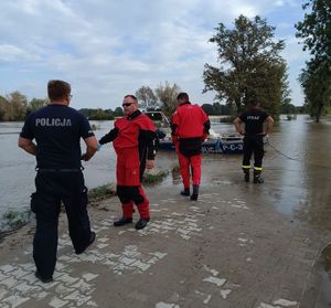 policjant ze strażakami nad wodą na której znajduje się policyjna łódka