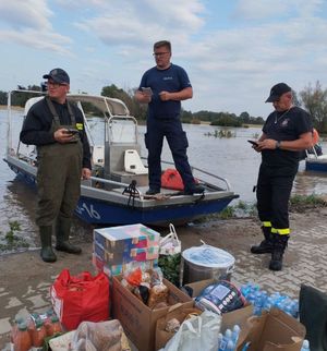 policjant stoi na łódce trzyma kartkę w dłoni obok dwóch strażaków i pudła z żywnością