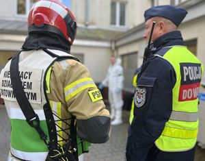 policjant dowódca strażak kierujący działaniami w tle osoba w białym kombinezonie