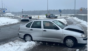 srebrny seat z rozbitym przodem stojący na chodniku przy rondzie w tle radiowóz i policjant