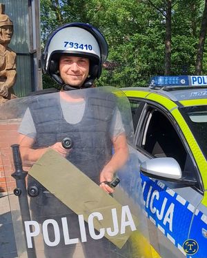 mężczyzna w hełmie z tarczą policja w tle radiowóz