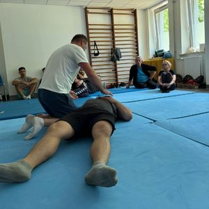 ćwiczenia na macie jeden mężczyzna leży drugi się nad nim pochyla w tle inni uczestnicy