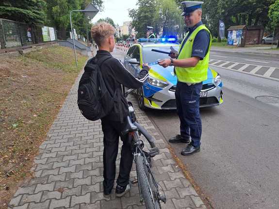 policjant ruchu drogowego rozmawiający z młodym chłopakiem na rowerze w tle radiowóz