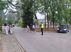 dzielnicowy stojący na skrzyżowaniu koło szkoły z prawej strony nadjeżdża samochód z lewej idą ludzie