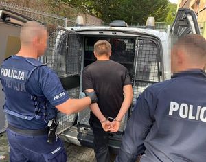 policjanci wprowadzający mężczyznę w kajdankach do zakratowanego radiowozu