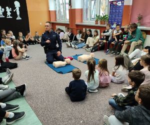 dzieci siedzące w kręgu pośrodku policjant pokazujący na fantomach pierwszą pomoc przedmedyczną