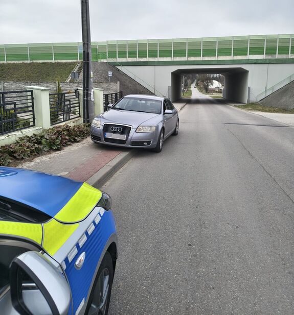 szare audi stojąca częściowo na chodniku przed nim widać przód radiowozu w tle wiadukt nad drogą
